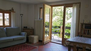 A cozy living room with a sofa, a small table, a radiator, and a sliding glass door leading to an outdoor area with a chair and a table. - Casa Lina by Interhome (Bondo)