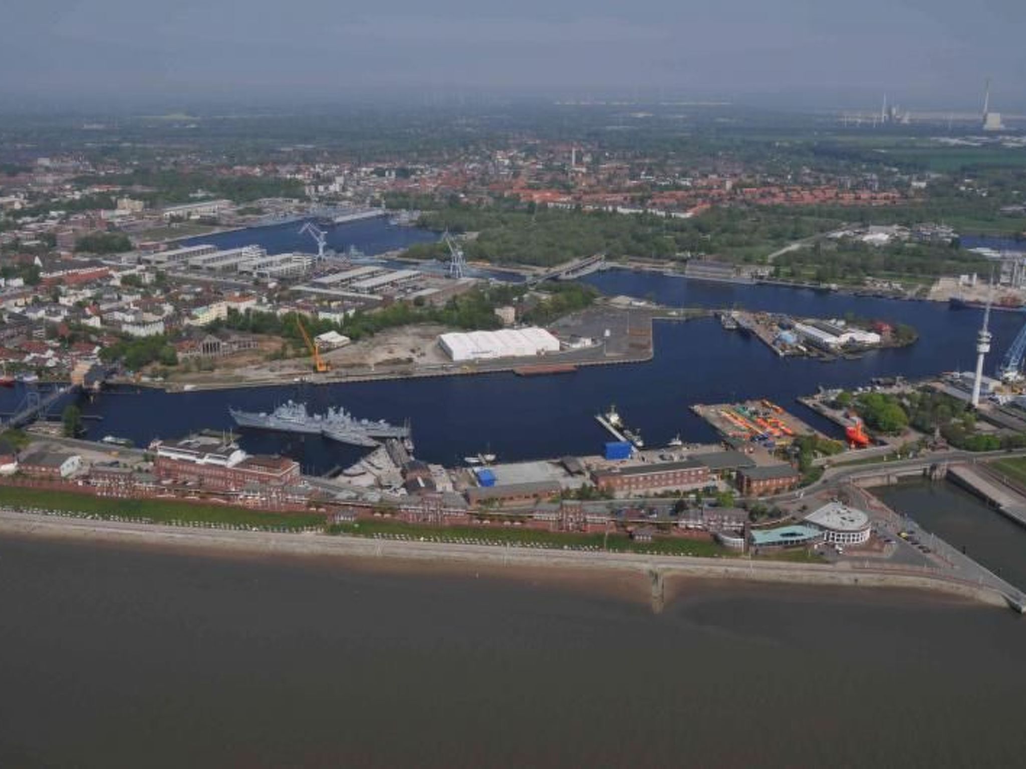An aerial view of a waterfront area with a large ship docked at a port, surrounded by industrial buildings and a bridge.