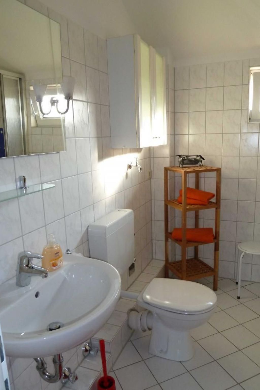 Une salle de bain avec un lavabo, des toilettes et un miroir, tous de couleur blanche. On y trouve une étagère en bois avec des serviettes orange et un porte-rouleau de papier toilette blanc.