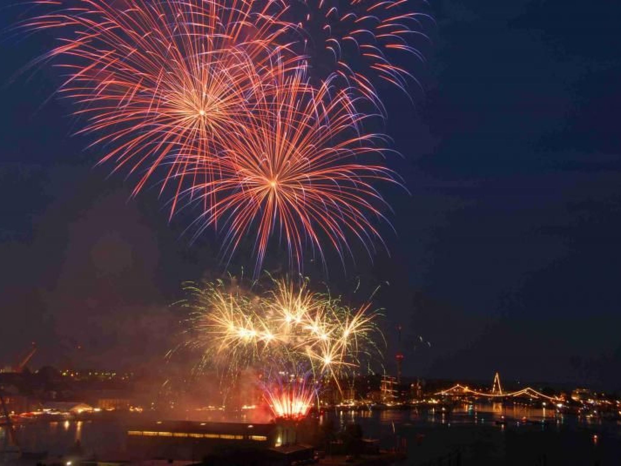 A nighttime view of fireworks over a waterfront with illuminated buildings and a bridge.
