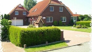 Une maison à deux étages, construite en briques, avec un toit recouvert de tuiles rouges, un garage et un jardin bien entretenu agrémenté de haies et d’une allée pavée.