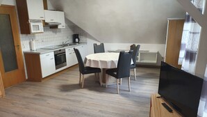 Une cuisine compacte avec des meubles blancs, un four à micro-ondes et une machine à café. On y trouve une table à manger avec quatre chaises et un téléviseur à écran plat posé sur un meuble en bois.