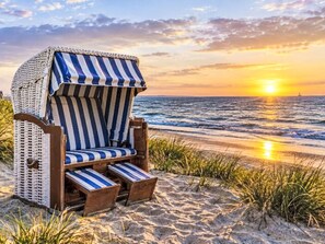 Ein Liegestuhl mit Streifenmuster, platziert an einem sandigen Strand, im Vordergrund Gras und im Hintergrund ein Sonnenuntergang über dem Meer.