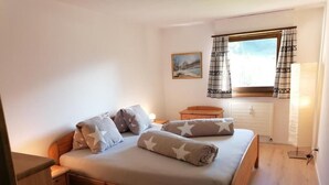 A bedroom with a wooden bed, a nightstand, a bedside table, a window with curtains, and a painting on the wall.