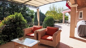 Area patio esterna con mobili in vimini, tavolo con piano in vetro e zona coperta con barbecue.