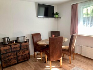 Ein Essbereich mit einem runden Tisch, vier Stühlen, einem Fernseher über dem Tisch, einer kleinen Ablage mit einer Pflanze, einer Kaffeemaschine und einem Toaster auf einem Holzschrank.