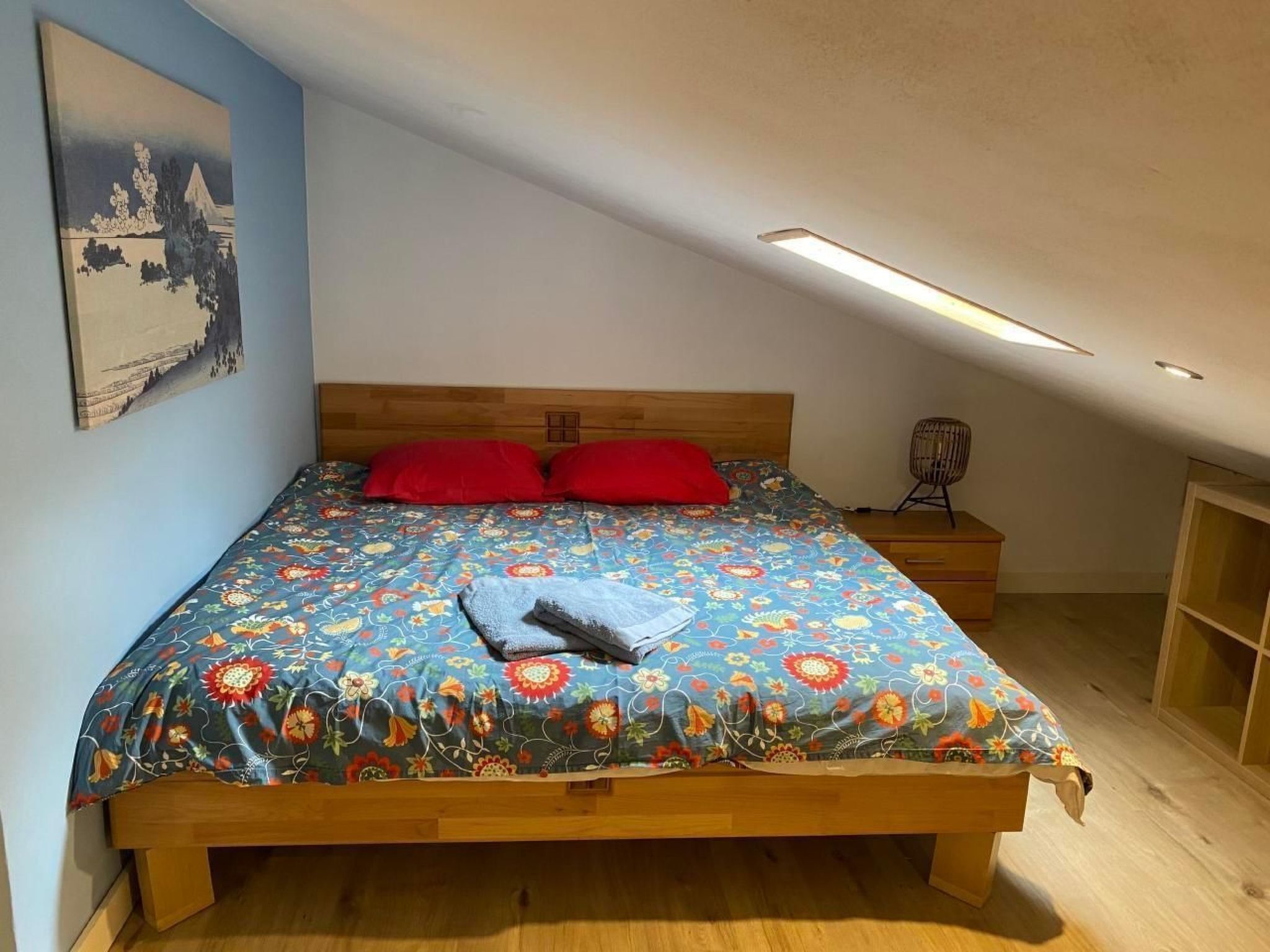 Een slaapkamer met een houten bed, een kleurrijk bloemenbeddengoed, rode kussens, een nachtkastje en een wandplank.