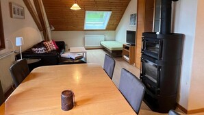 Ein gemütliches Zimmer mit Holzbalkendecke, einem Esstisch, einer Couch, einem Fernseher und einem Kaminofen.