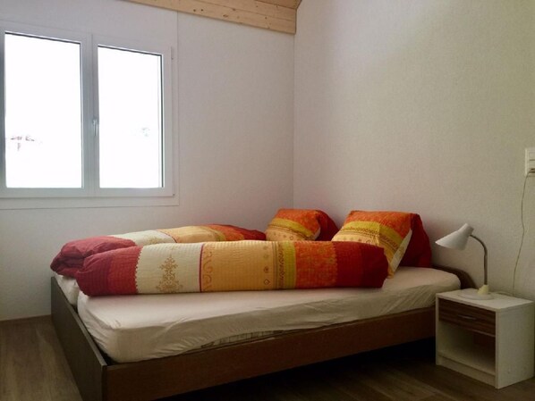 A single bed with a wooden frame, white bedding, and a colorful blanket. There is a nightstand with a lamp, a window with blinds, and a white wall.