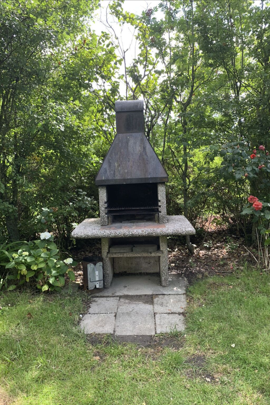 Un barbecue extérieur en pierre, doté d’une cheminée, entouré d’une végétation luxuriante et de plantes en fleurs.