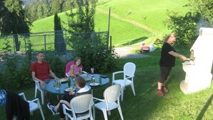 Un groupe de personnes est assis autour d’une table ronde à l’extérieur, des chaises sont disposées autour de la table, et celle-ci est mise pour le repas.