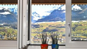 A window with a view of a mountainous landscape, a green wall, and two potted plants. - Seeblick by Interhome (Sarnen)