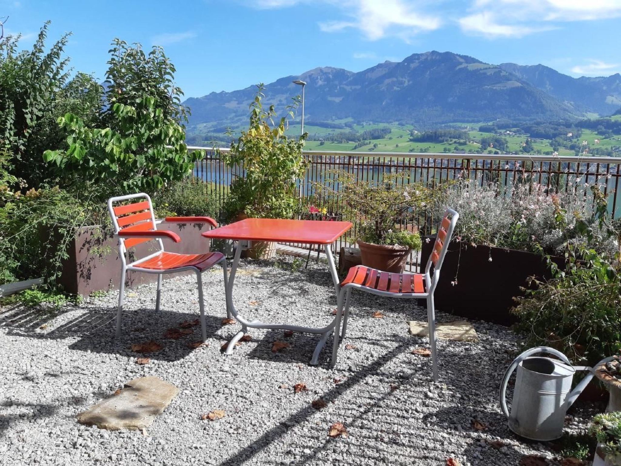 Une terrasse avec deux chaises rouges et une table, entourée de gravier, d’où l’on peut admirer une vue sur les montagnes et un pont en arrière-plan.