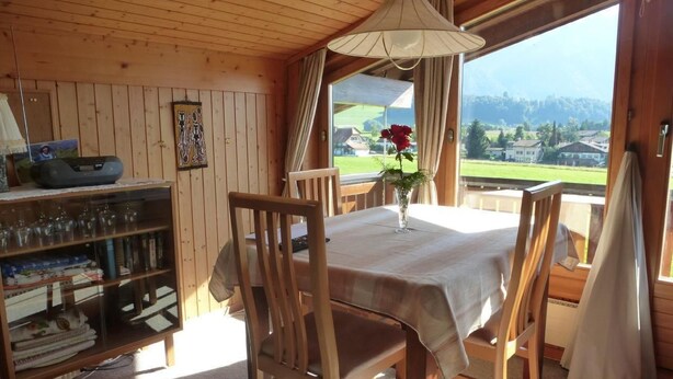 Un interior de madera con una mesa de comedor, sillas, un florero con flores y una estantería con vasos y libros.