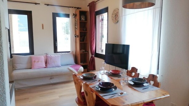Un comedor con una mesa de madera dispuesta para dos personas, un televisor en la pared y un sofá al fondo.