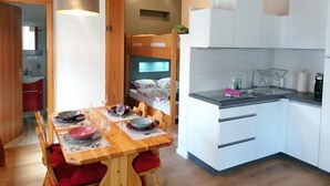 Cocina compacta con armarios blancos, una mesa de madera para cuatro comensales y una cama litera al fondo.
