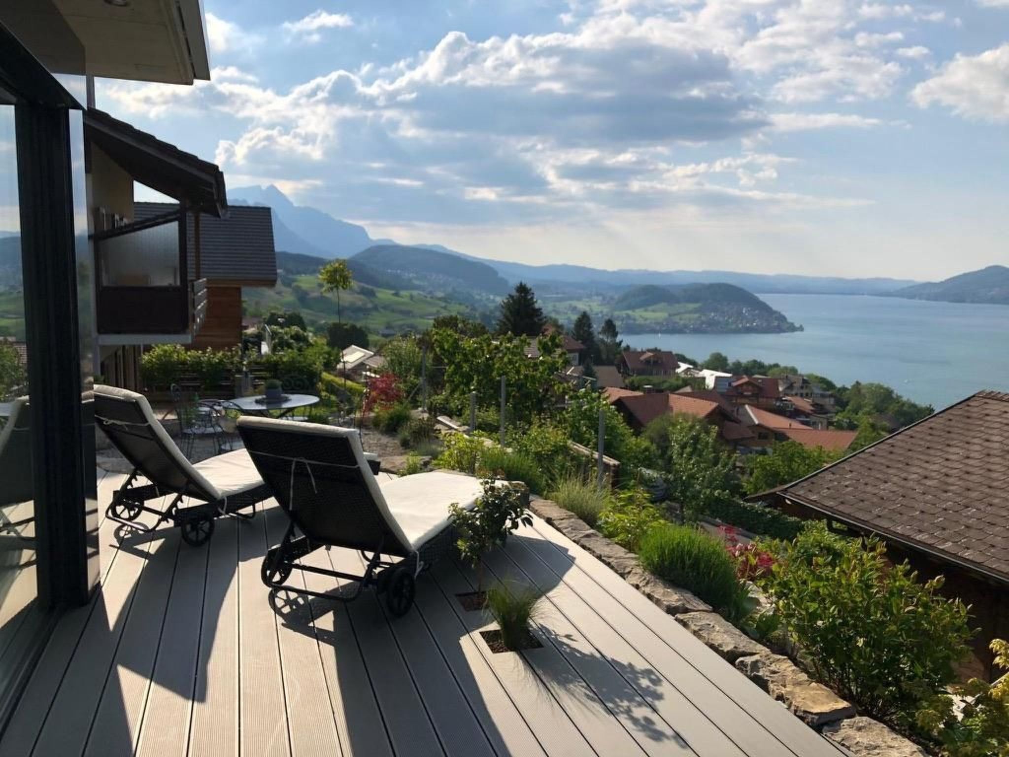 A deck with two lounge chairs and a table, overlooking a lake and a village.