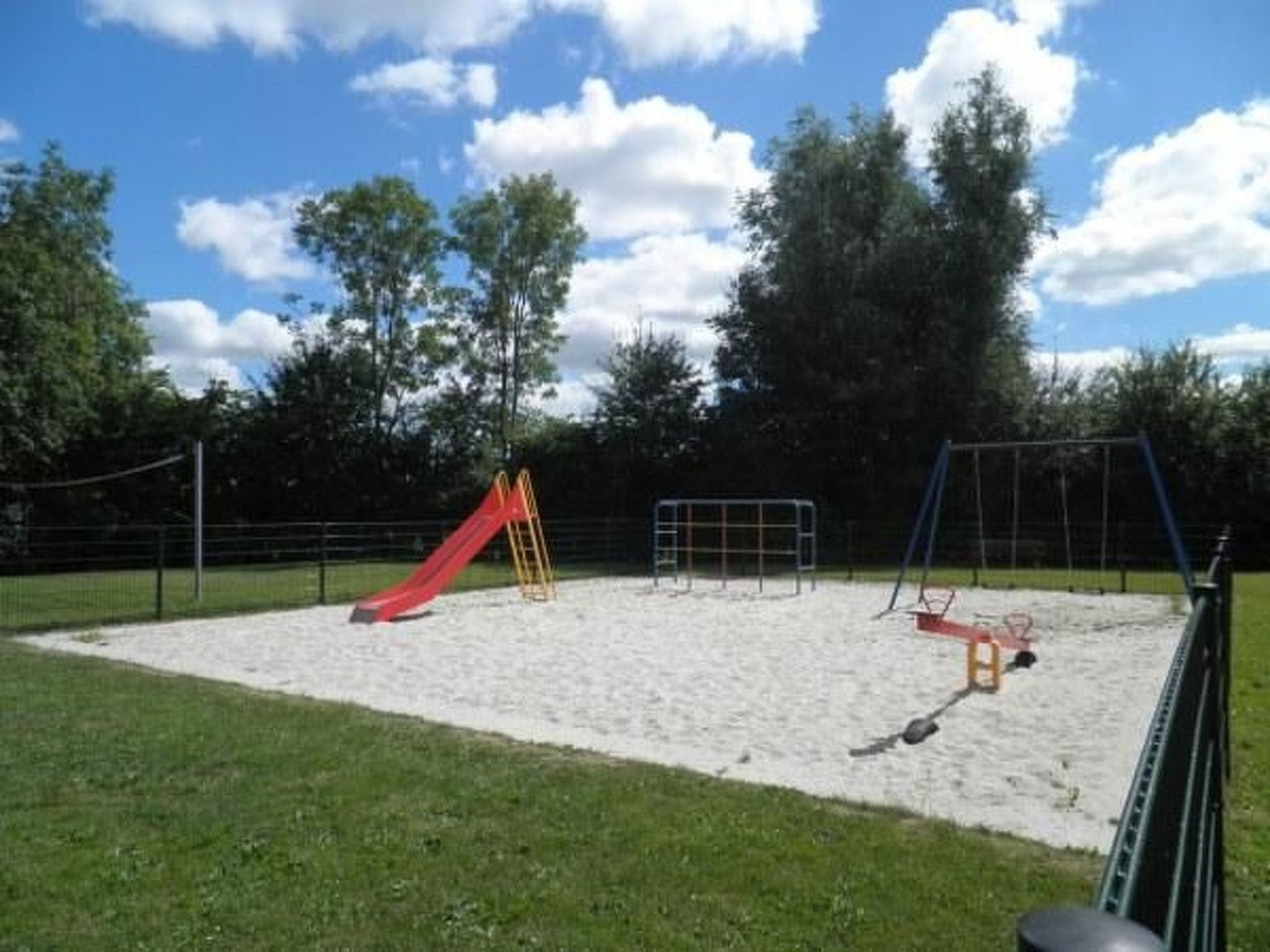 Un parque infantil con un tobogán rojo y columpios, rodeado de una valla y árboles.