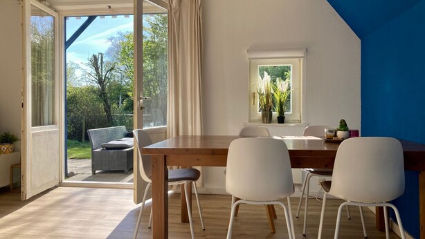 A dining area with a wooden table and white chairs, a window with plants, and a view of greenery outside. - 162 Grachthaus by Interhome (Anjum)