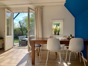Een eethoek met een houten tafel en witte stoelen, een raam met planten, en uitzicht op groen.