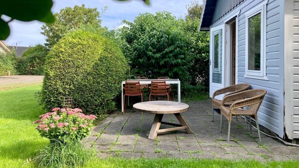 Eine Terrasse mit rundem Tisch und Stühlen, ein kleiner Garten mit rosa Blüten und ein Haus mit weißer Fassade und einem Fenster.