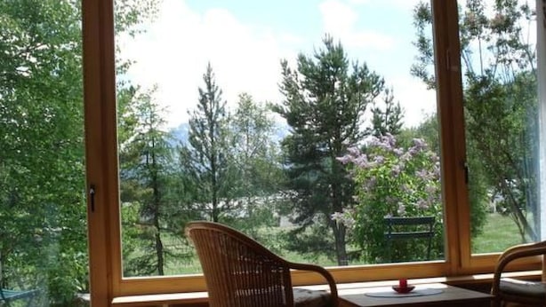 Una stanza con una grande finestra che si affaccia su alberi e una montagna. Si vede un tavolo di legno e due sedie in vimini.