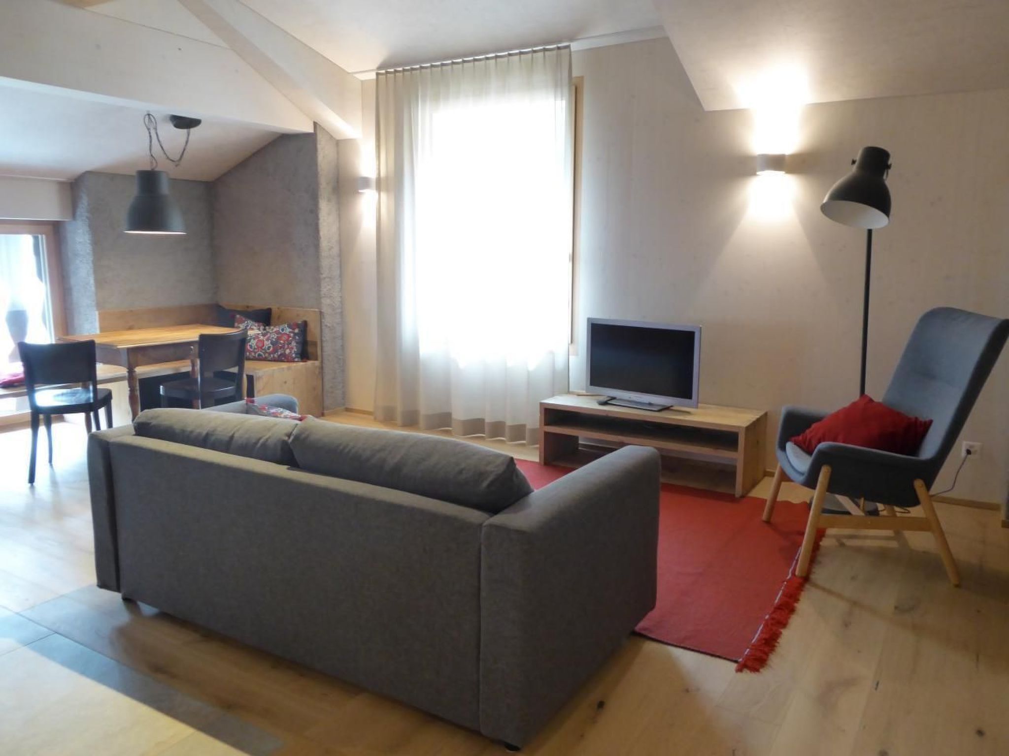 Un salon avec un canapé gris, un téléviseur posé sur un meuble en bois, un tapis rouge, un fauteuil bleu et un coin repas avec une table et des chaises en bois.