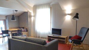 Una sala de estar con un sofá gris, un televisor sobre un mueble de madera, un tapete rojo, un sillón azul y una zona de comedor con una mesa y sillas de madera.