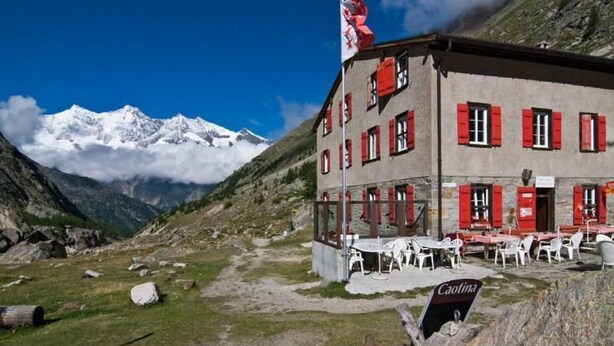 A building with red shutters and a flag, surrounded by a mountainous landscape and a clear sky. - Haus Mondelli by Interhome (Saas-Almagell)