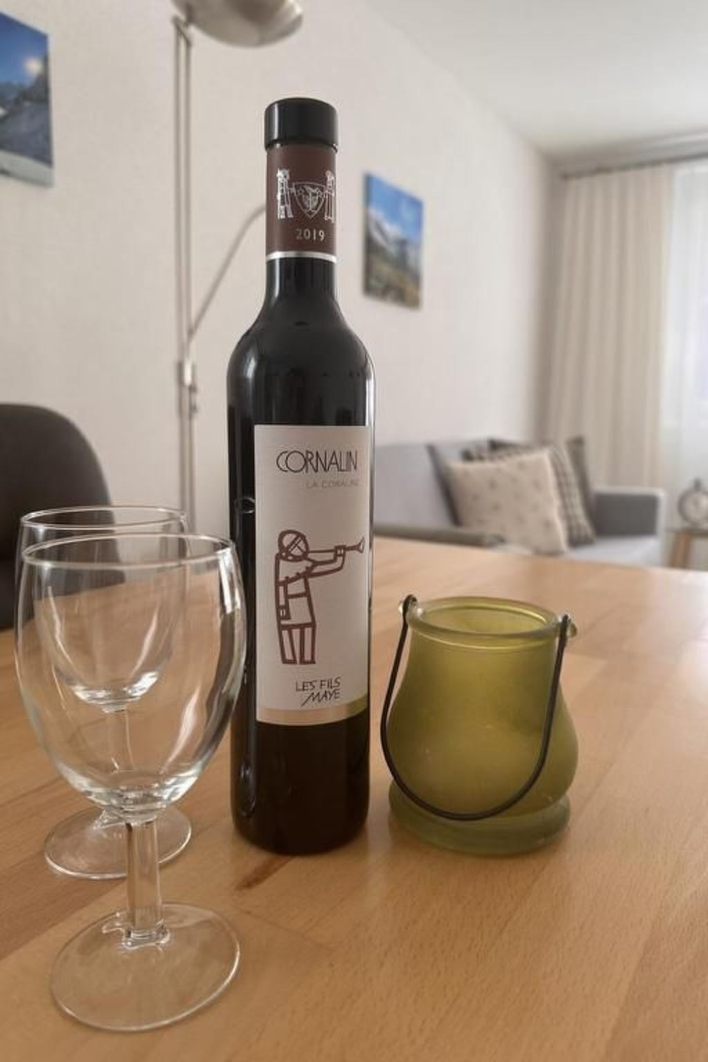 Une bouteille de vin Cornalin, un verre et une bougie sur une table en bois, dans une pièce éclairée par une lampe et comportant un canapé.