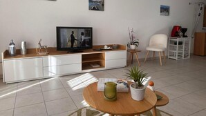 Ein modernes Wohnzimmer mit einem Flachbildfernseher, einem Holztisch, einem weißen Sessel und einem kleinen Beistelltisch mit einer roten Kaffeemaschine.