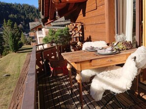Una terraza de madera al aire libre con una mesa de comedor y sillas blancas mullidas, rodeada por una valla de madera y vistas a la montaña.