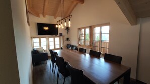 Ein geräumiger Essbereich mit einem langen Holztisch und mehreren Stühlen, einem Wand-Fernseher und großen Fenstern für natürliches Licht.