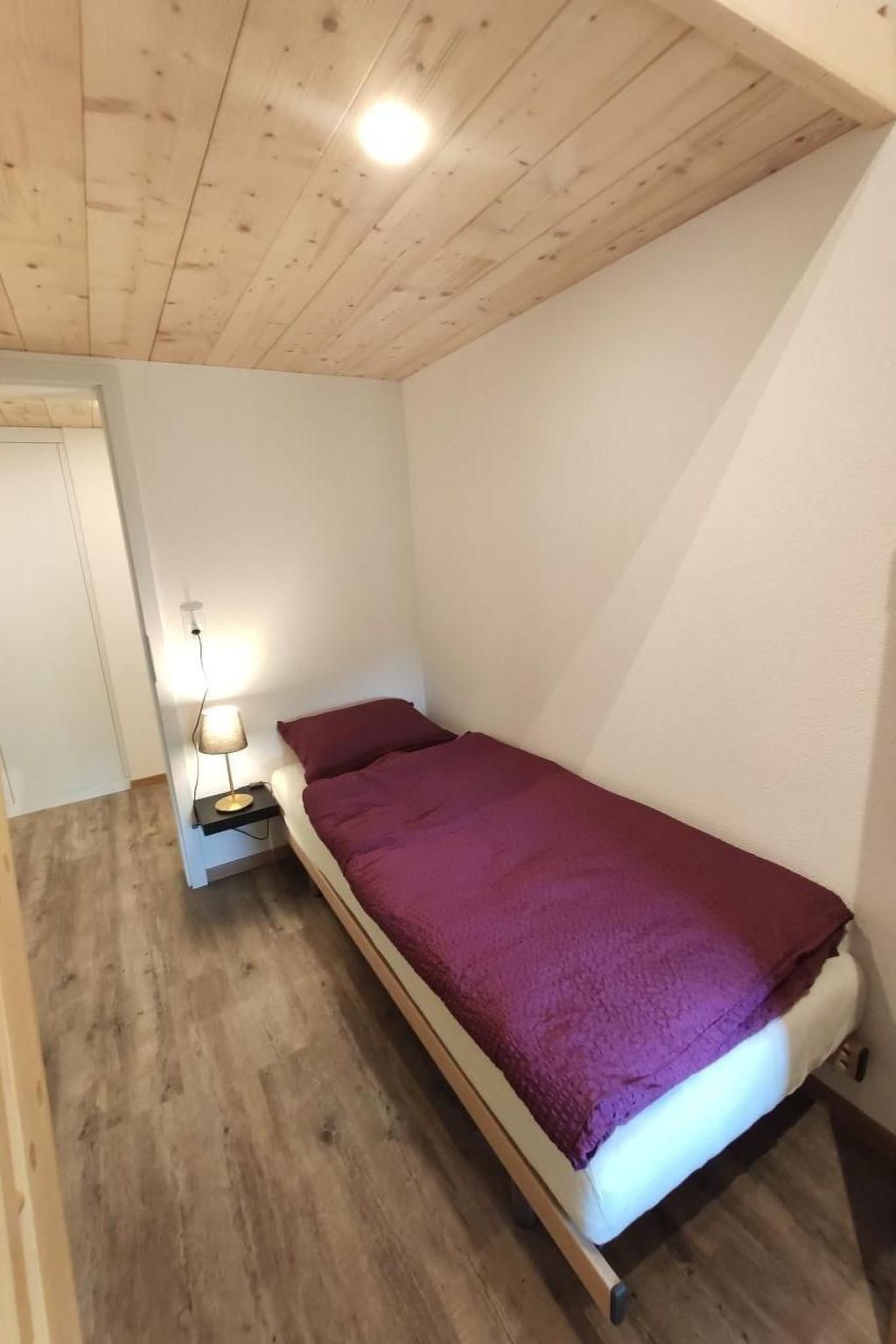 Una piccola camera con un letto singolo, soffitto in legno e comodino con lampada.
