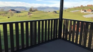 Un balcón de madera con vista a un paisaje rural, que incluye casas y colinas.