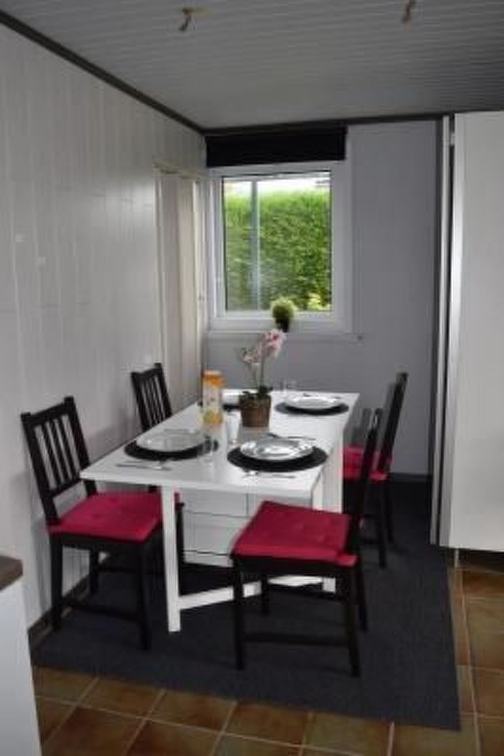 A dining area with a white table set for four, black chairs with red cushions, a window with a view of greenery, and a tiled floor.