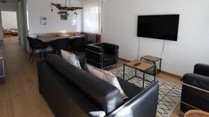A modern living room with a black leather sofa, a dining area with a wooden table and chairs, a flat-screen TV mounted on the wall, and a unique ceiling light fixture. - Siesta 3,5-Zimmer-Wohnung by Interhome (Leukerbad)