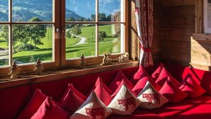 Habitación con un ventanal grande que ofrece vistas a un paisaje verde y montañas. Hay cojines rojos en un sofá rojo, y un estante pequeño con figuritas.