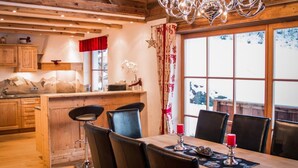 Interior acogedor de una cabaña con una mesa de comedor de madera, sillas negras, una araña y un ventanal con vistas a árboles cubiertos de nieve.