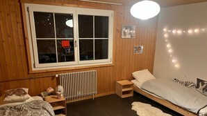 A cozy bedroom with a wooden floor, a bed with a grey blanket, a small bedside table with a lamp, a radiator, and a window with curtains. - mit Bergblick by Interhome (Rueras Dieni)