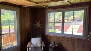 A cozy room with wooden walls and a sloped ceiling, featuring a large window with a view of a village, a wooden chair with a fur throw, and a star decoration on the wall. - mit Bergblick by Interhome (Rueras Dieni)