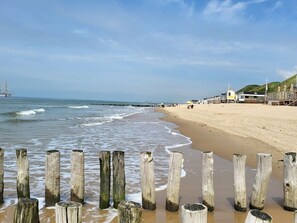 Una playa con postes de madera en primer plano, una orilla arenosa y un muelle que se adentra en el agua.