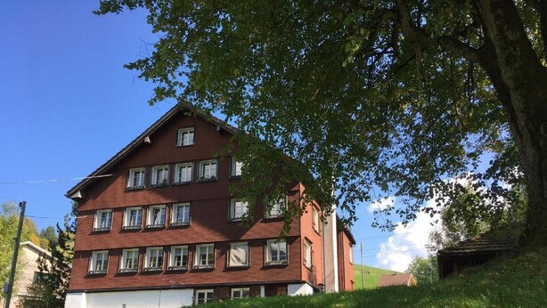 En byggnad med flera våningar, med en röd fasad och vita detaljer, omgiven av frodig grönska och en klarblå himmel.