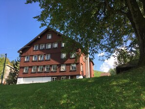 Ein mehrgeschossiges Gebäude mit roter Fassade und weißer Ornierung, umgeben von üppigem Grün und blauem Himmel.
