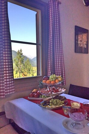 Ein Esstisch mit Obst- und Käseplatte, einem Dessertglas und Blick auf Berge durch ein Fenster.
