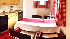 Una cocina compacta con zona de comedor, que incluye una mesa con mantel blanco, sillas, una lámpara colgante y cortinas a cuadros.