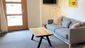 Un salon moderne avec un canapé gris, une table basse en bois, un téléviseur à écran plat fixé au mur et un tableau abstrait aux couleurs vives.