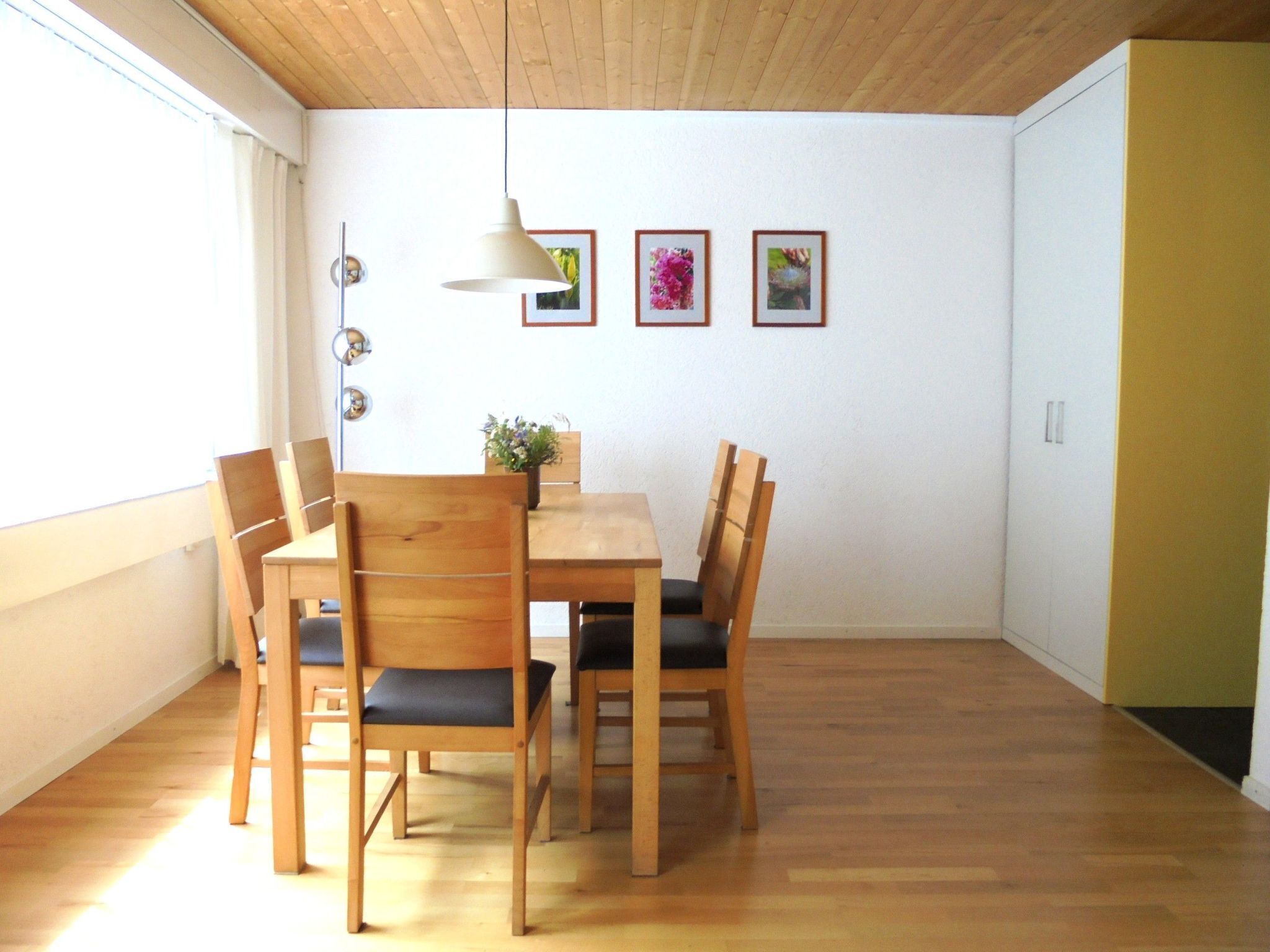 A dining area with a wooden table and chairs, a hanging light fixture, and framed pictures on the wall.