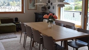 Un'area pranzo con tavolo in legno, sedie e un vaso di fiori. Due finestre offrono vista su una zona boschiva.