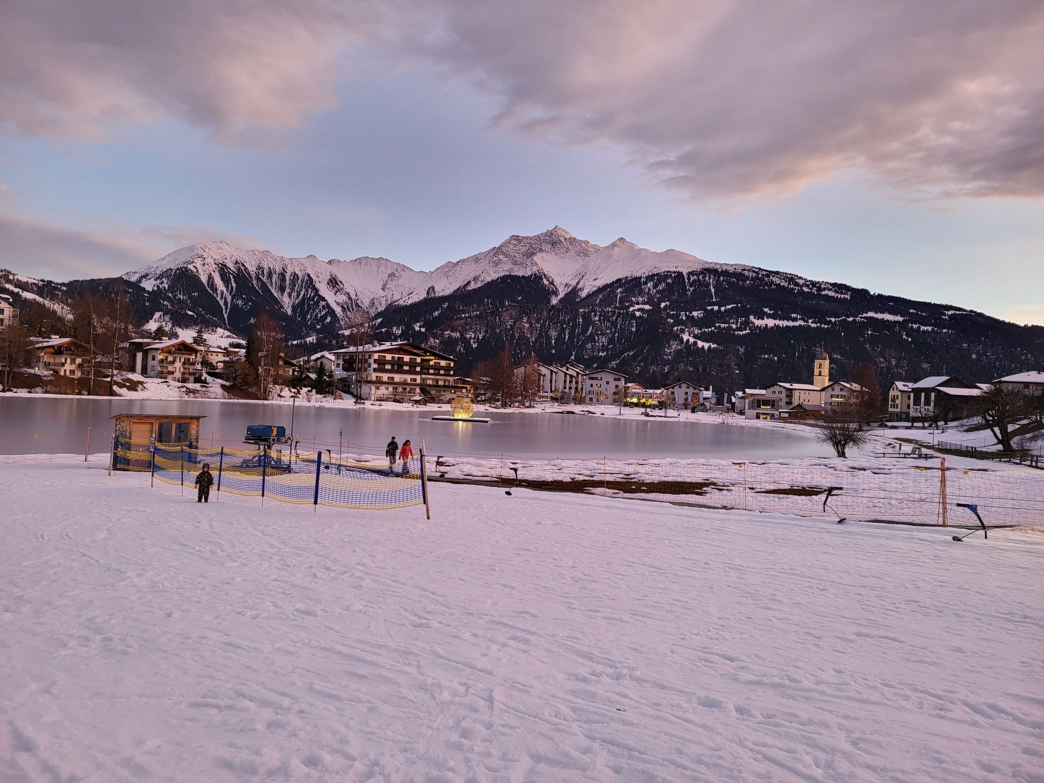 Un lac recouvert de neige, avec des bâtiments et des montagnes en arrière-plan.